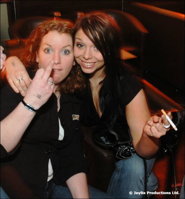 Two women pose on a sofa, one making a rude gesture and the other holding a cigarette