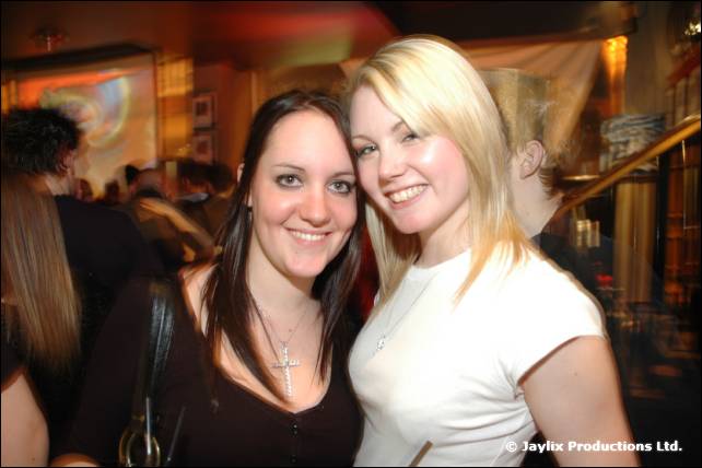 Two smiling women stand close together in a busy bar, one with blonde hair in a white top