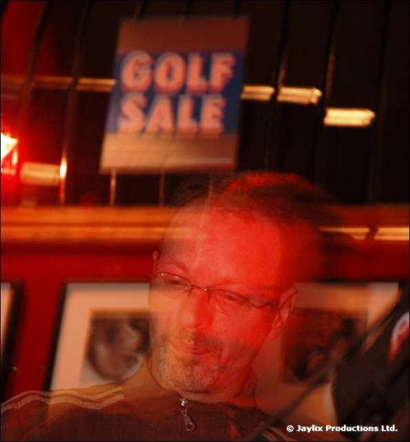 A blurred man in glasses sits below a blue Golf Sale sign in a red room