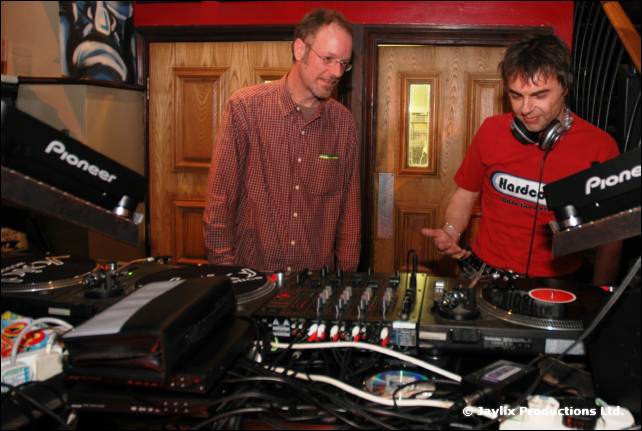 Two men at DJ decks between Pioneer mixers, one in a checked shirt, one in a red t-shirt wearing headphones