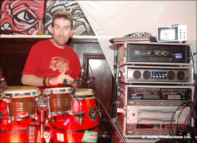 A man in a red t-shirt plays congas beside a rack of audio equipment and a small television