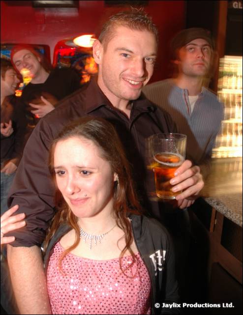 A man holding a pint stands behind a woman in a pink sparkly top, looking unhappy, at a bar