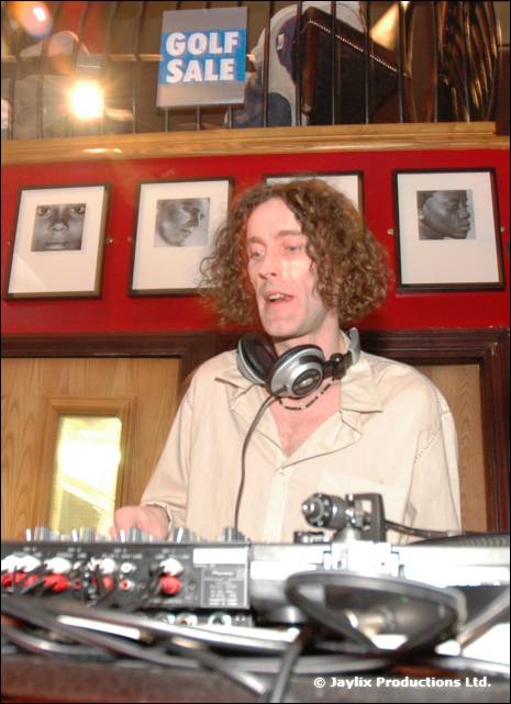 A man with curly hair and headphones sings or plays at a DJ mixer beside framed pictures