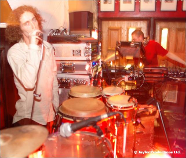 A man with curly hair sings into a microphone beside congas, with another person at decks behind