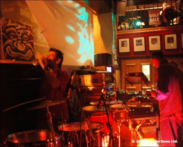 Musicians perform on a small stage with drums and a projected cyan pattern behind