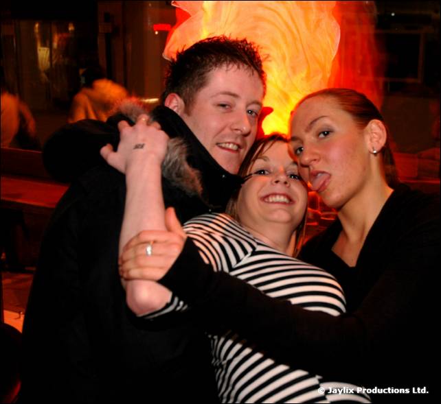 Two people and a third person with their tongue out pose close together in a bar, with an orange glow behind