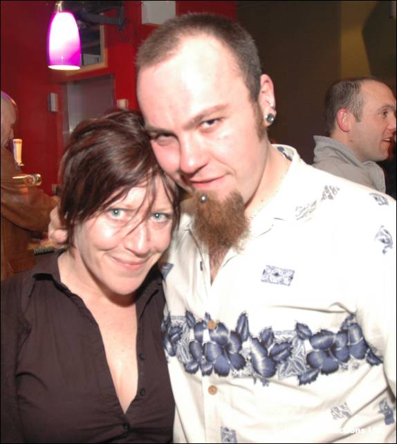A woman with short dark hair and a man with a goatee in a patterned shirt pose close together in a bar