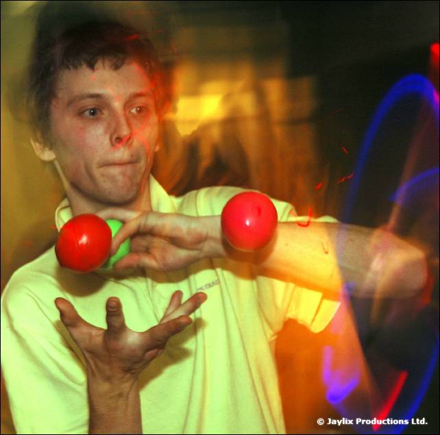 A person in a pale green top juggles two red balls, with blue light trails behind