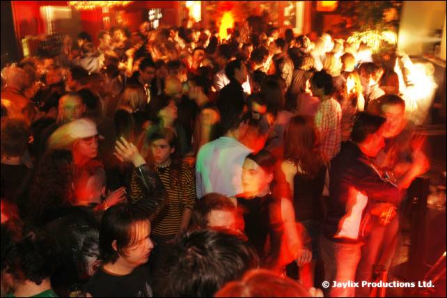 A dense crowd fills a dimly lit bar under red and orange lighting