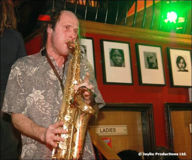 A man in a patterned shirt plays a saxophone beside framed portraits and a Ladies sign