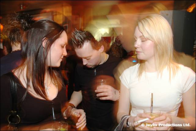 Two women and a man with spiky hair stand close together holding drinks, with motion blur