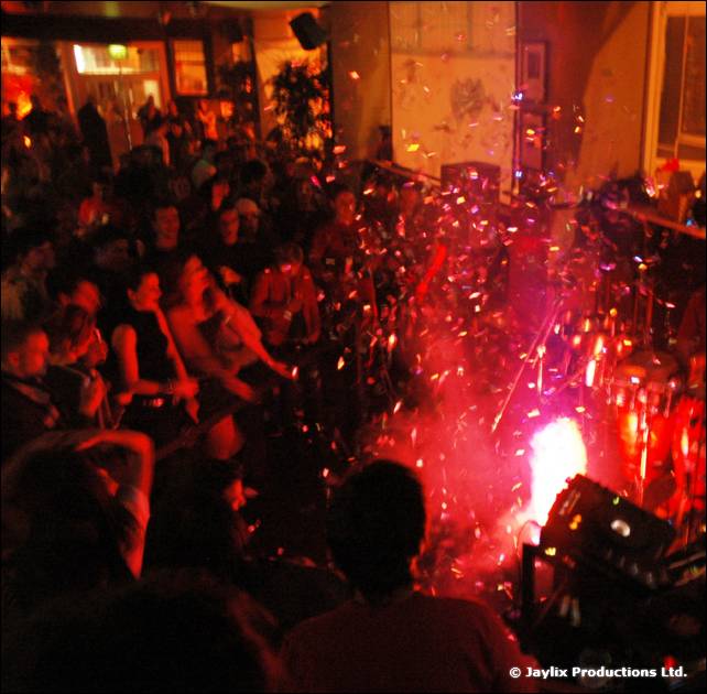 A dark crowd watches sparks and a bright flare near the DJ equipment, lit in red