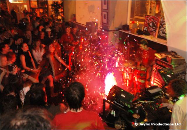 A crowd gathers around DJ decks as sparks and glittering confetti fill the air under red light