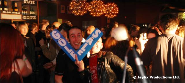 A smiling person holds up a blue foam arrow reading Golf Sale in a crowded bar