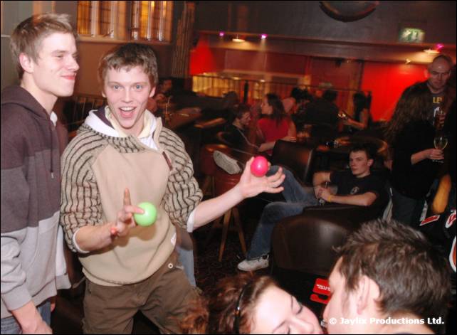 Two young men stand in a bar, one holding a pink ball and a green ball, with people seated behind