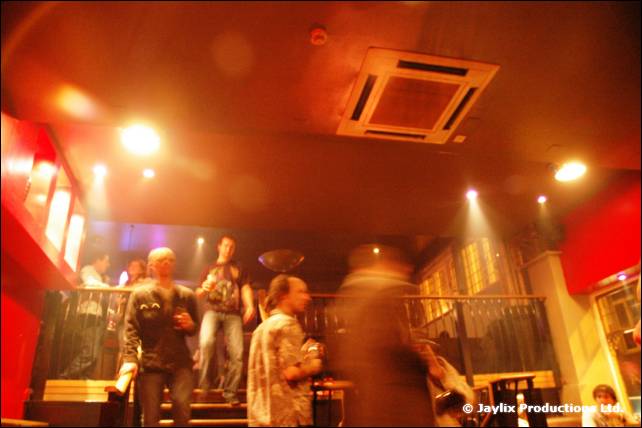 A quiet bar interior with a few people standing near a stair rail, lit in orange
