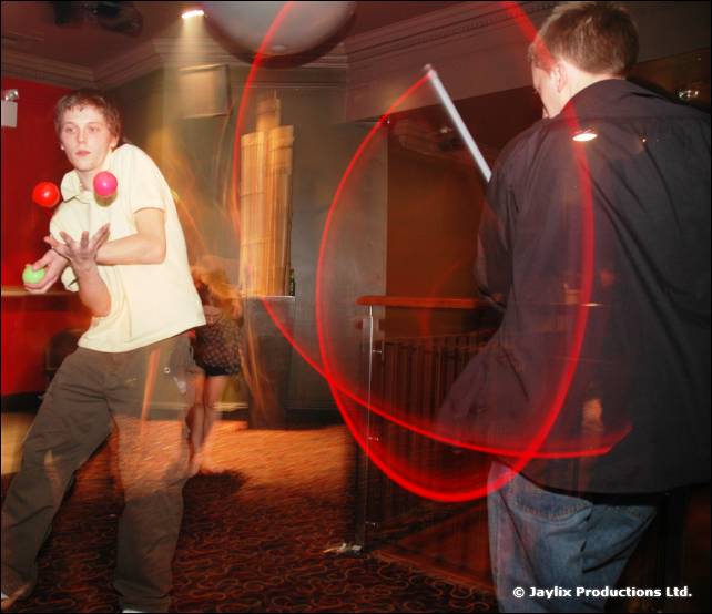 A person juggles two balls while another person in a dark jacket swings a staff leaving a red light trail