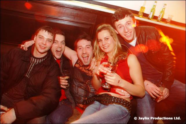 Five people sit close together on a banquette, smiling at the camera, with a woman in red in the middle