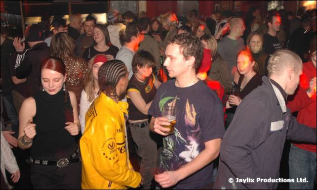 A crowd on a dance floor, with a person in a yellow jacket and a man holding a drink in the foreground