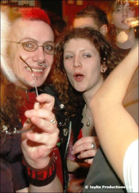 A person with a red mohican and glasses holds up a glowing stick beside a woman with curly hair