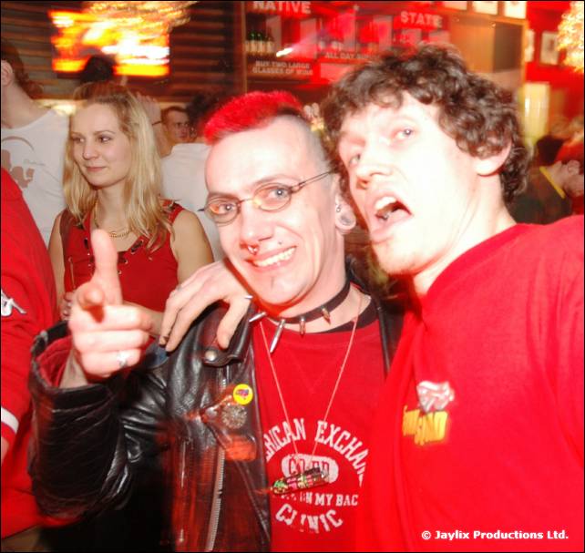 A person with a red mohican and glasses and a curly haired man in a red top pose, one pointing at the camera