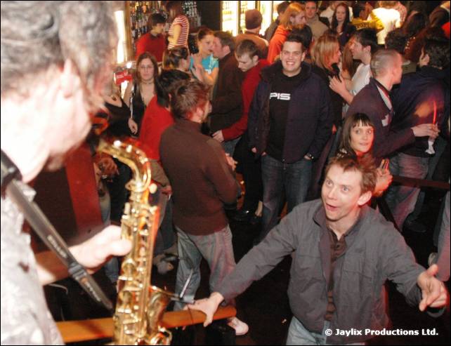 A saxophone in the foreground as a man in a grey jacket gestures with open arms in a crowd