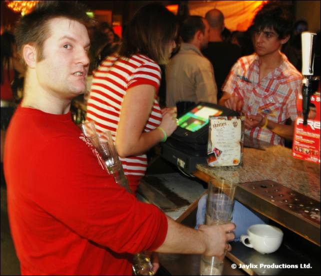 A man in a red top stands at a bar holding a glass, with other people and a till behind