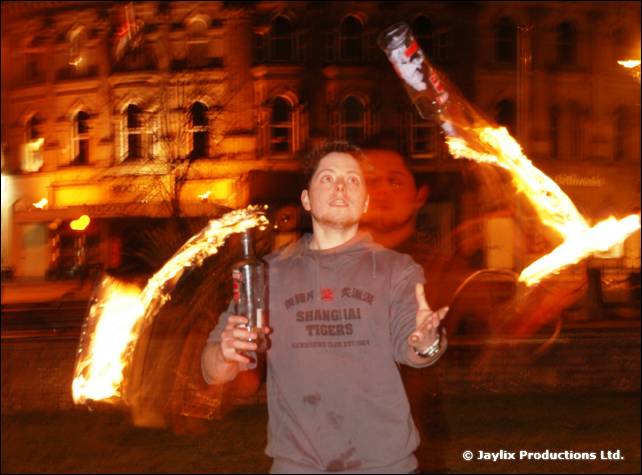 A person in a grey Shanghai Tigers sweatshirt holds a bottle with flames swirling around them at night