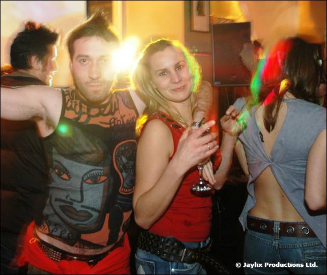A man in a patterned top and a woman in red hold a drink, with a bright flare behind them