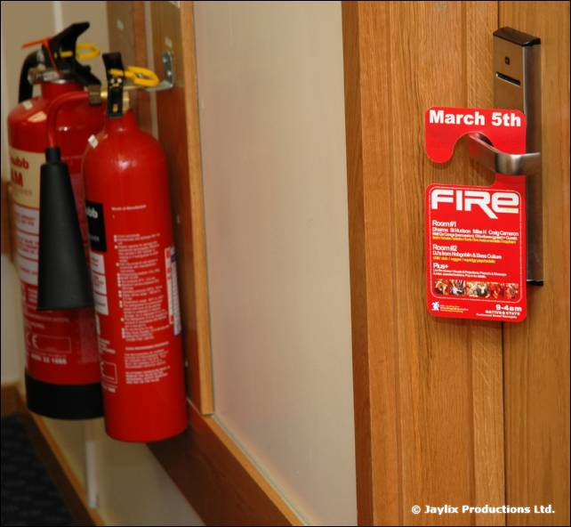 A red FIRE door hanger dated March 5th on a wooden door beside two red fire extinguishers