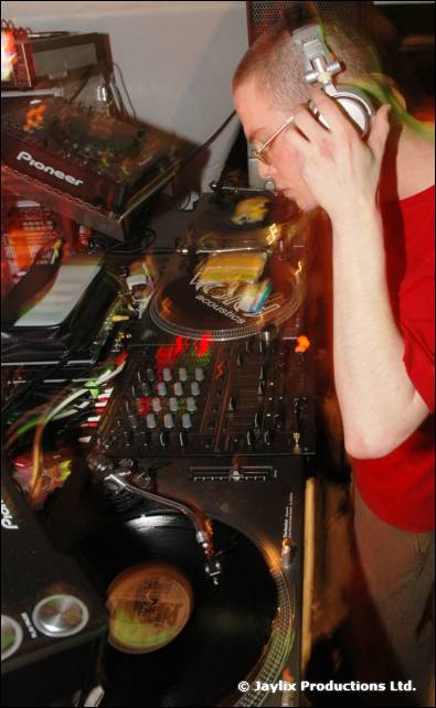 A DJ in headphones and a red top leans over Pioneer decks and a turntable