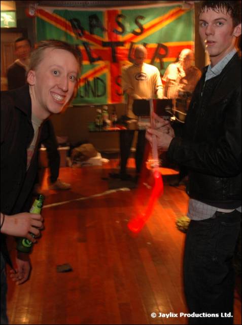 Two young men hold bottles in front of a Union Jack flag with painted lettering, with a red light trail