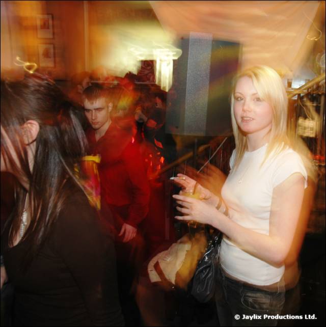 A woman in a white t-shirt holds a drink in a crowded bar, with blurred people beside her