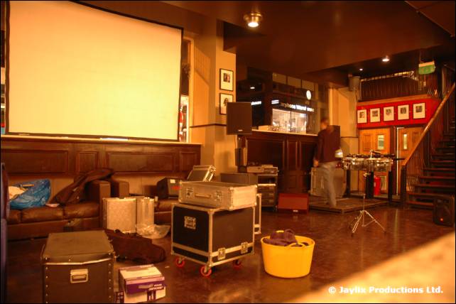 A blank projector screen above flight cases and a yellow tub in a bar being set up