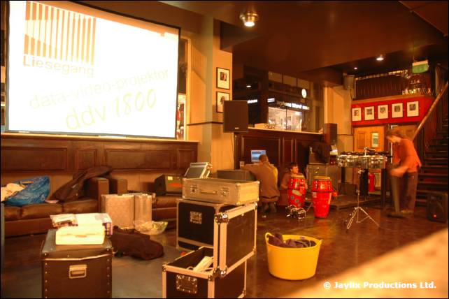 A projector screen showing a barcode and text above flight cases and a yellow tub in a bar
