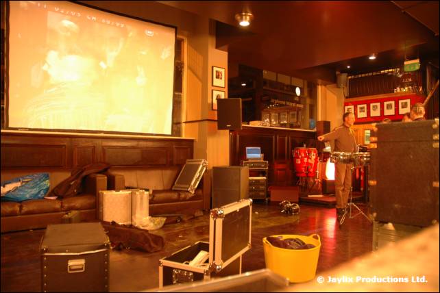 A projector screen shows a dim picture above flight cases in a bar, with a person setting up percussion