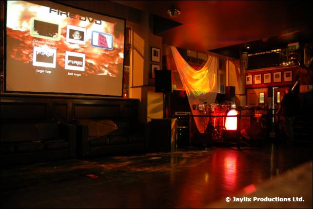 A projector screen showing a FIRE DVD menu in a dark bar, with red lights and draped fabric beside it