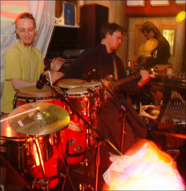Two musicians play drums and a guitar on stage near a cymbal, lit in orange