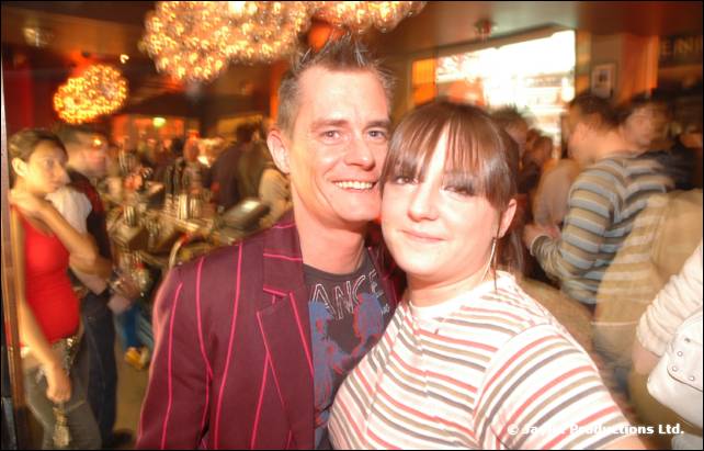 A man in a striped jacket and a woman in a striped top pose close together in a busy bar