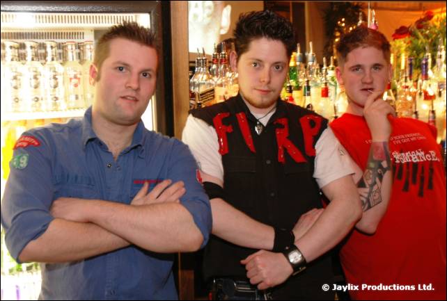 Three men in front of a bar stand with arms folded, one wearing a black FIRE waistcoat