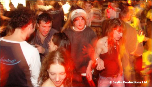 A crowd dances in a busy bar under orange light, with blurred movement