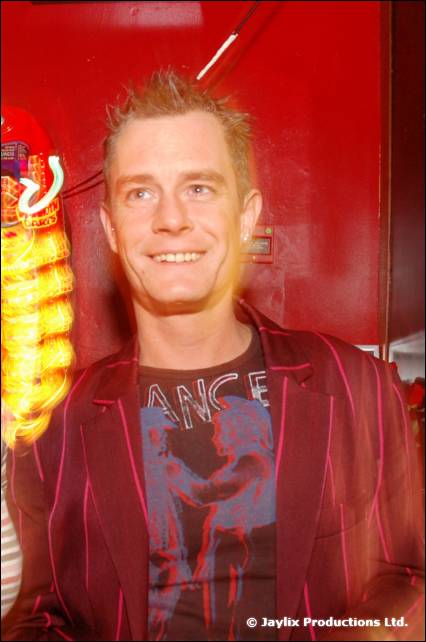 A smiling man in a pinstriped blazer and printed t-shirt against a red wall, with a streak of light beside him