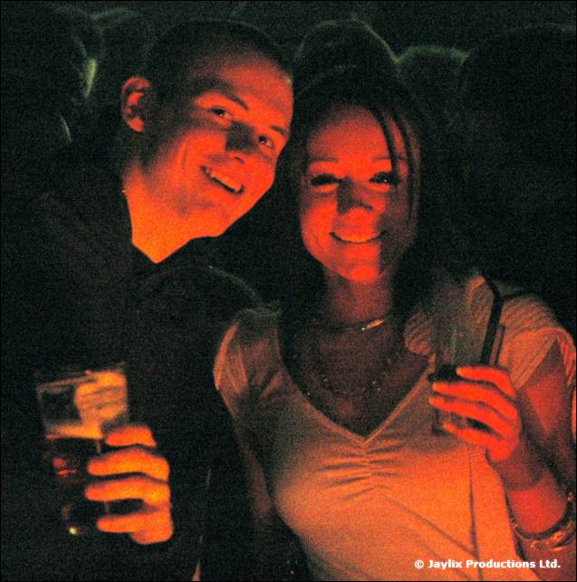A smiling man and woman hold drinks in a dark room lit in red