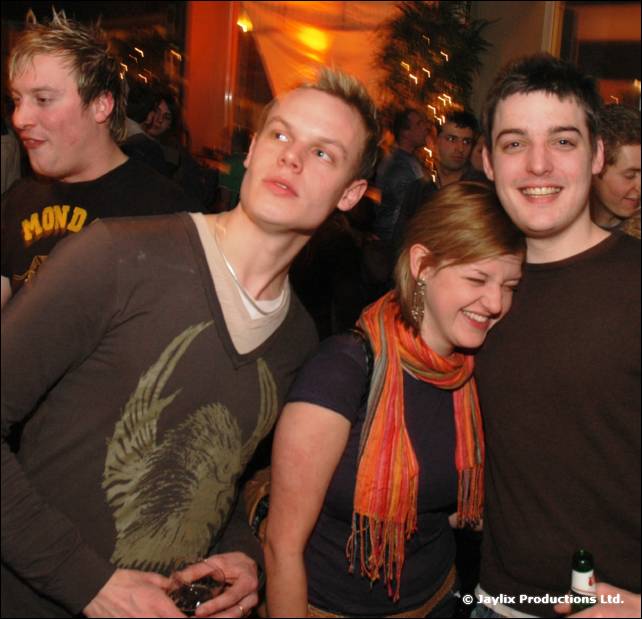 Two men and a woman with an orange scarf smile together in a crowd, one man holding a bottle
