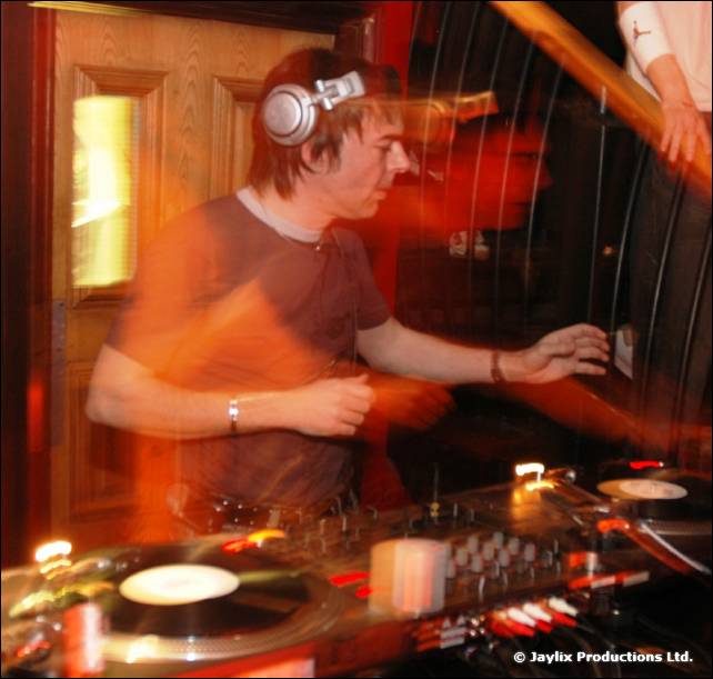 A DJ in headphones mixes at turntables in front of a wooden door, blurred with motion