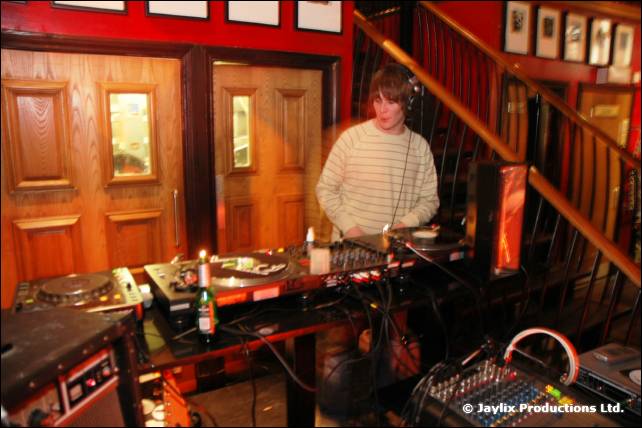 A DJ in a striped top stands at decks and a mixer beside a wooden staircase