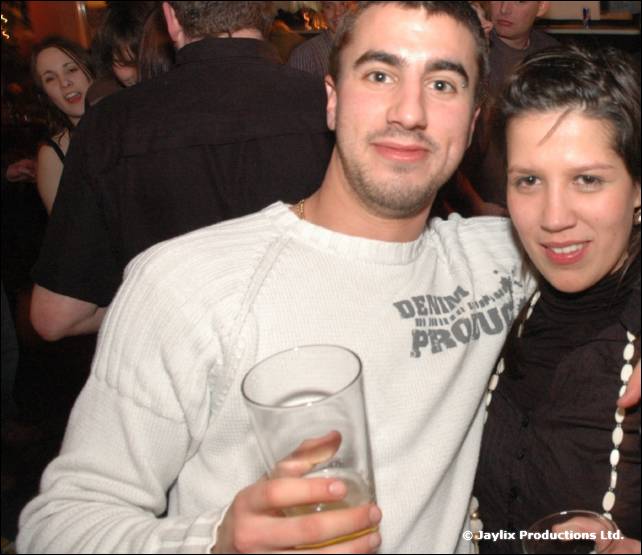 A smiling man in a white sweatshirt holds a pint glass beside a woman with short dark hair