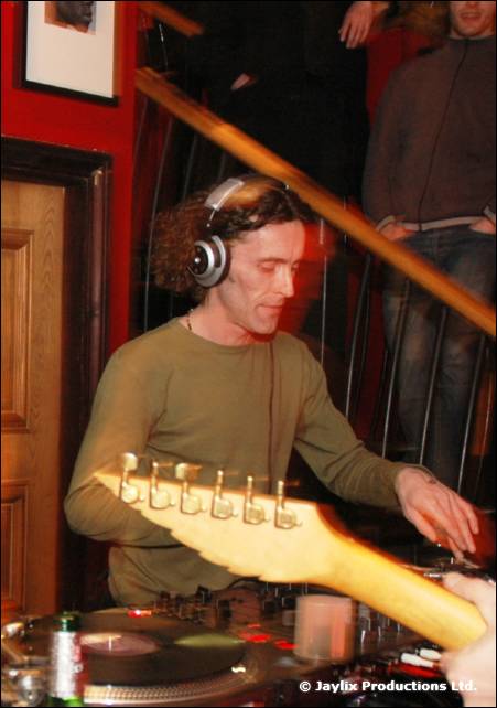 A DJ in headphones and a green jumper at decks, with the neck of a guitar across the frame