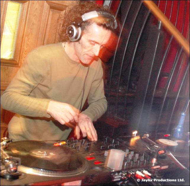 A DJ with curly hair and headphones mixes at a turntable and mixer, wearing a green jumper
