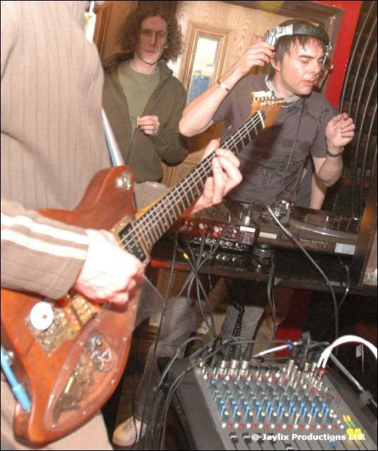 A man plays an electric guitar beside a mixing desk while a person in headphones stands behind